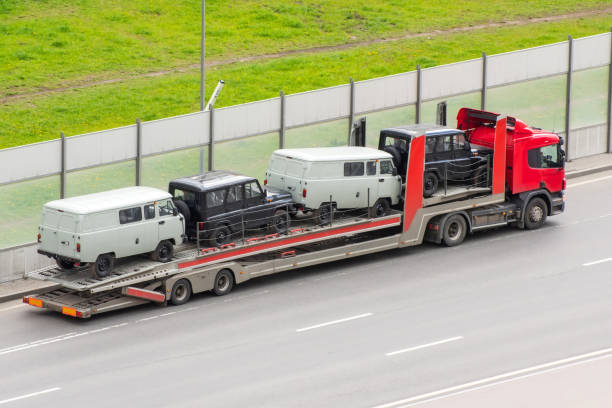 A red car carrier truck from MD Auto Transport Gaithersburg transports three vintage vans—two white and one black—along a road with a grassy area and a sound barrier in the background.