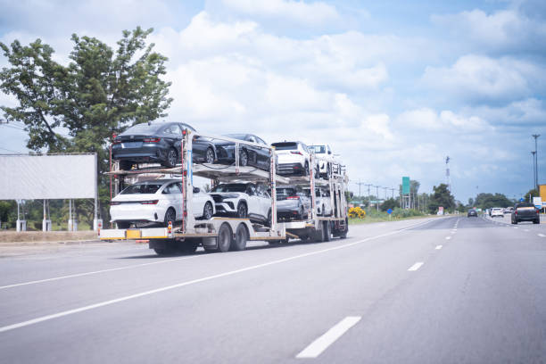 A car carrier truck loaded with multiple vehicles, part of an MD Car Shipping Gaithersburg operation, drives down a highway under a partly cloudy sky, surrounded by trees and road signs.