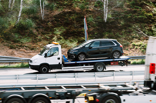 Tow truck carrying a damaged vehicle on its flatbed while driving on a highway, illustrating roadside assistance and vehicle recovery services.